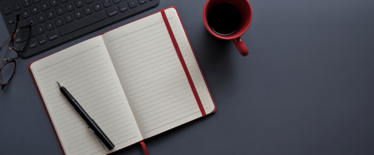 Carnet vide ouvert avec un stylo noir, une tasse de café et un clavier à proximité, symbolisant le début de l’écriture d’un roman sans idée claire.