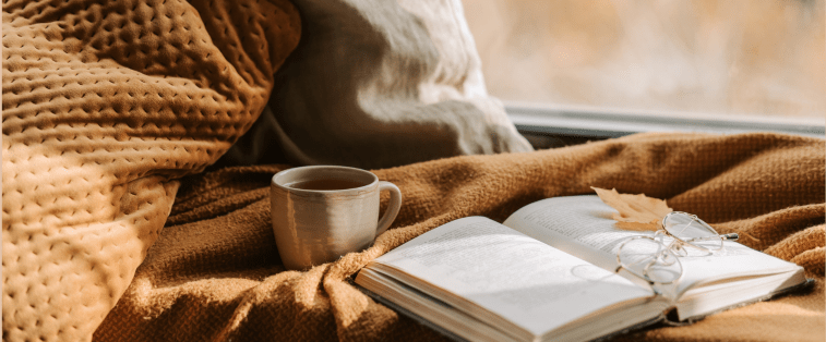Livre ouvert sur une couverture en laine avec une tasse de thé et des lunettes, posé devant une fenêtre : une scène cosy et chaleureuse, parfaite pour relire ses romans préférés.