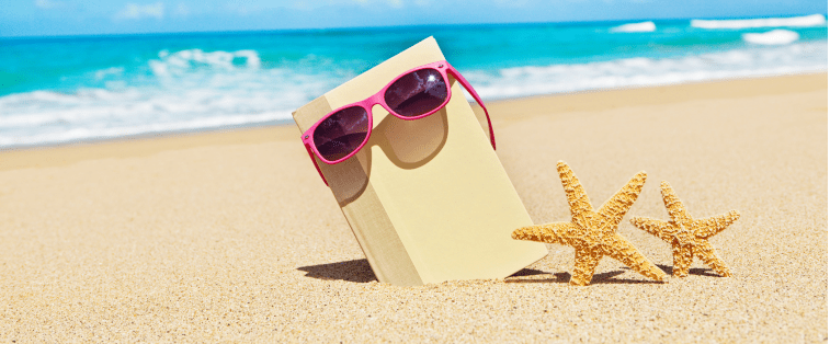 Livre posé sur le sable avec des lunettes roses et des étoiles de mer, prêt pour une lecture d’été au bord de la mer.