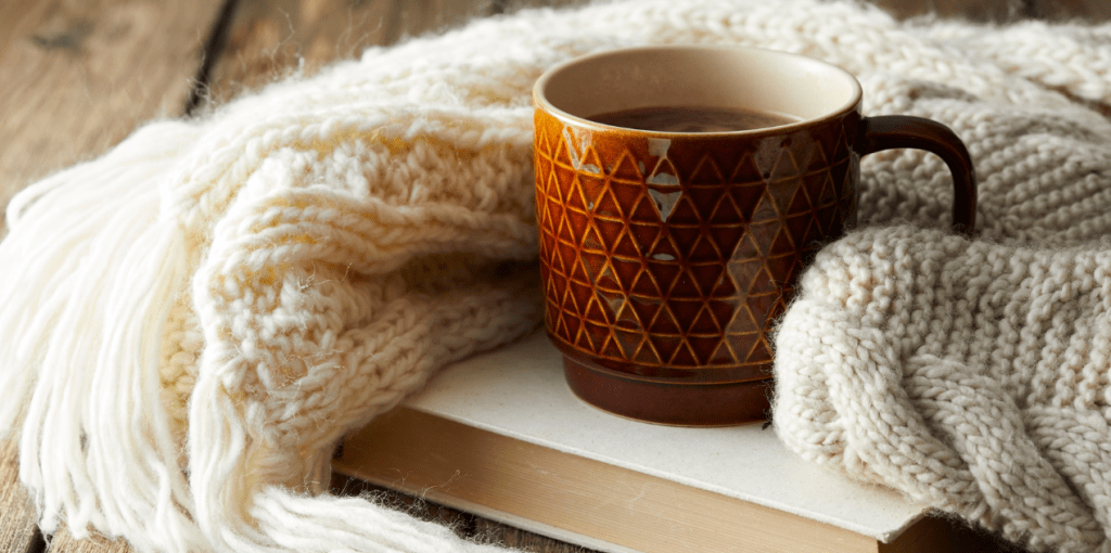 Tasse de chocolat chaud posée sur un livre avec un plaid en laine beige, ambiance cosy d’automne parfaite pour une lecture doudou.