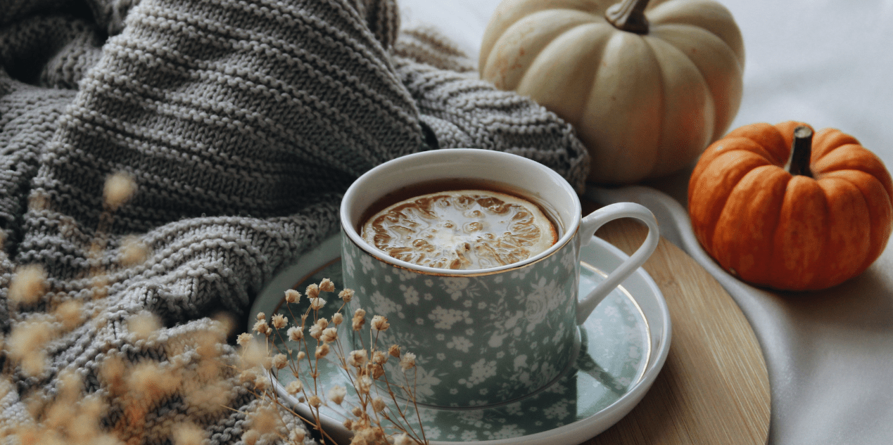 Tasse de thé aux agrumes posée sur une soucoupe fleurie, entourée de citrouilles décoratives et d’un plaid tricoté gris, ambiance automne cosy pour Halloween réconfortant.